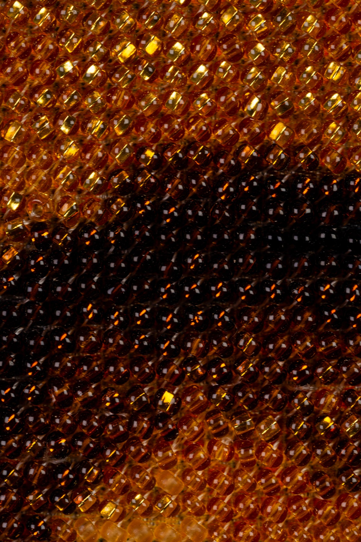 Extreme macro of amber, gold and dark brown seed beads revealing the dense hand-embroidered surface of the artwork